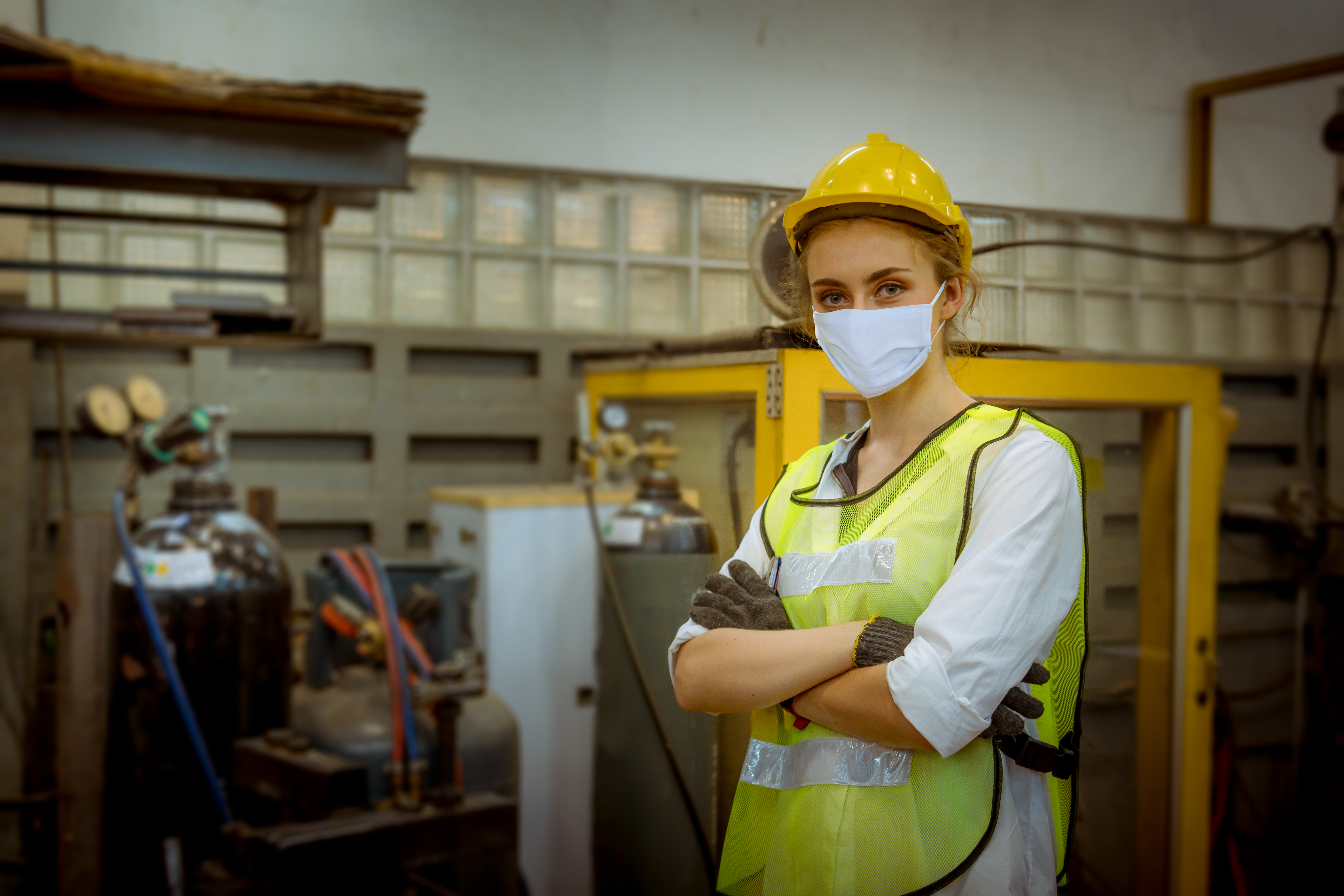A frontline worker stands with arms crossed wearing a yellow safety helmet, a medical mask, and a yellow safety vest. 